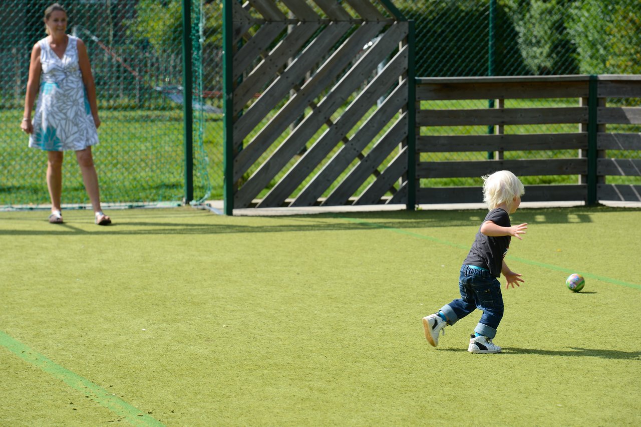 A young child runs toward a small ball on a playground while an adult watches from a distance.