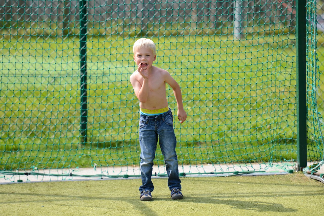 A shirtless young boy in jeans stands in front of a playground net, touching his face and looking ahead.