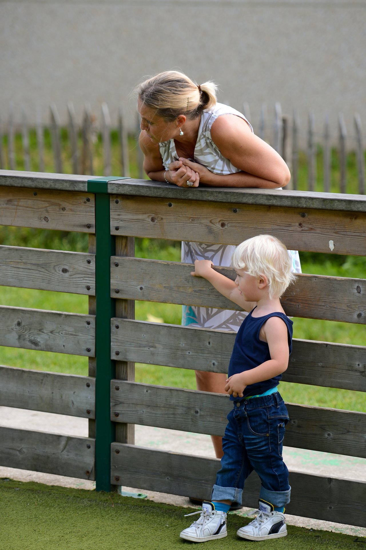 A young child points through a wooden fence while an adult leans over, watching and listening attentively.