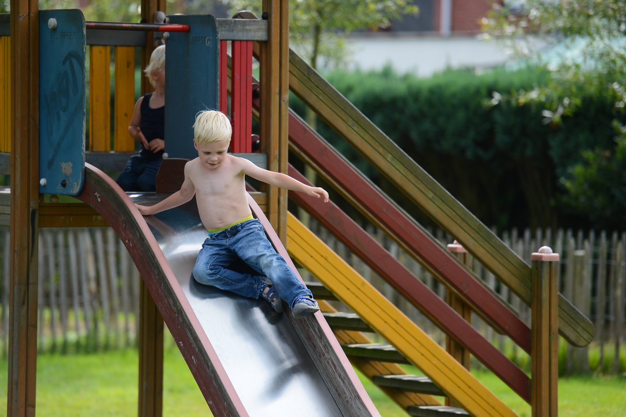 A shirtless child slides down a playground slide, while another child sits at the top watching.
