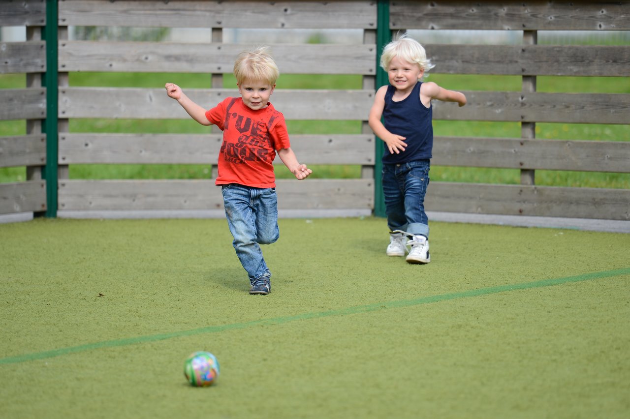 Two young children run on a playground chasing a colorful ball, smiling and playing together.