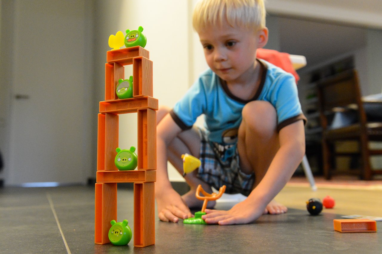 A young child prepares to launch a toy bird at a stacked tower with green pig figures.