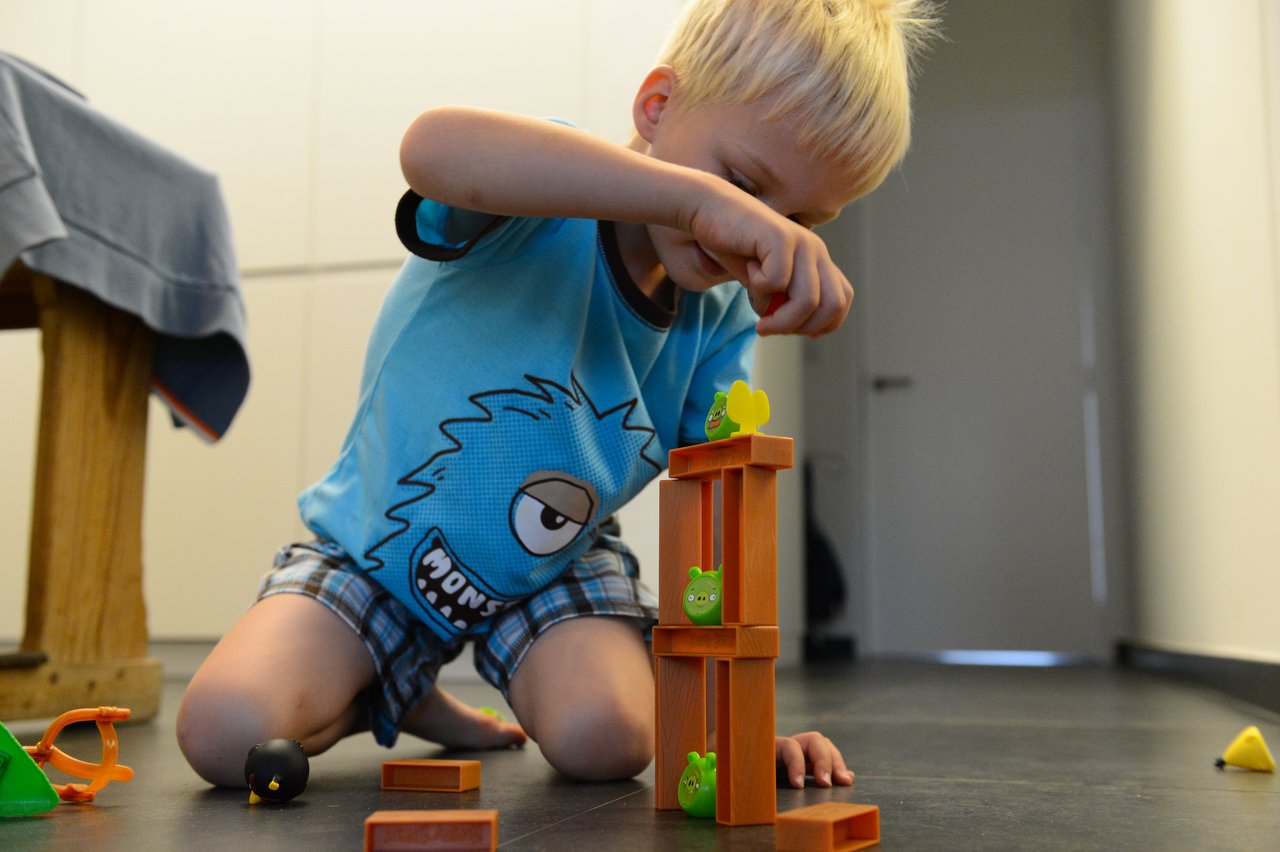 A child kneels on the floor, carefully stacking toy blocks and Angry Birds figures in a tower.