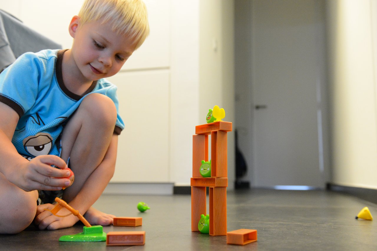 A young child plays with an Angry Birds toy set, launching a bird at a tower with green pig figures.