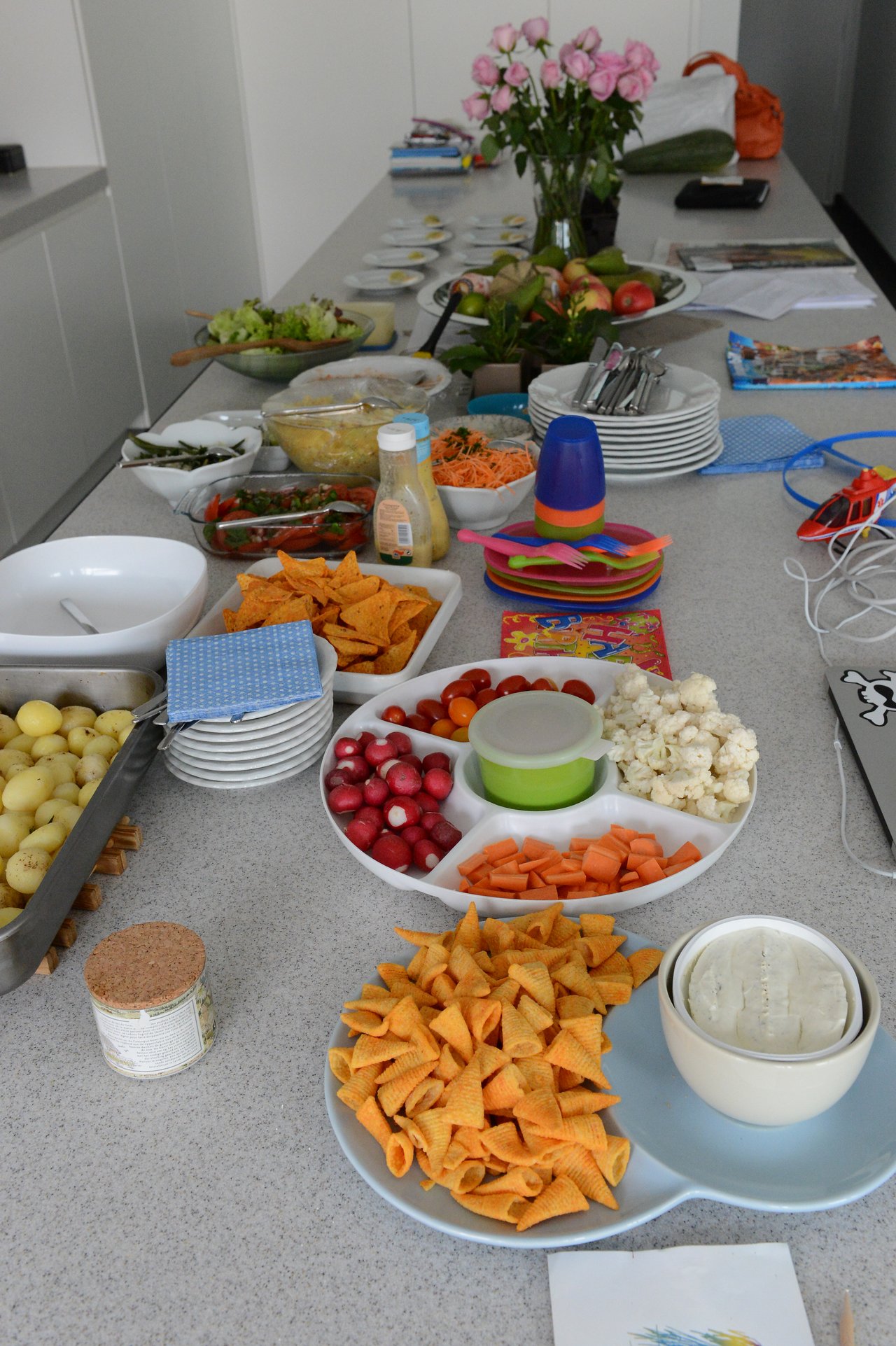 A table filled with snacks, vegetables, chips, and dips, prepared for a barbecue gathering.