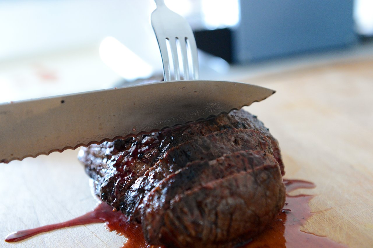 A knife and fork cut into a grilled steak on a wooden cutting board, with juices visible.