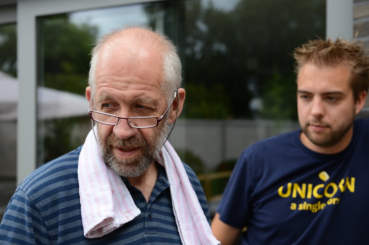 An older man with glasses and a towel on his shoulder stands next to a younger man in a t-shirt.