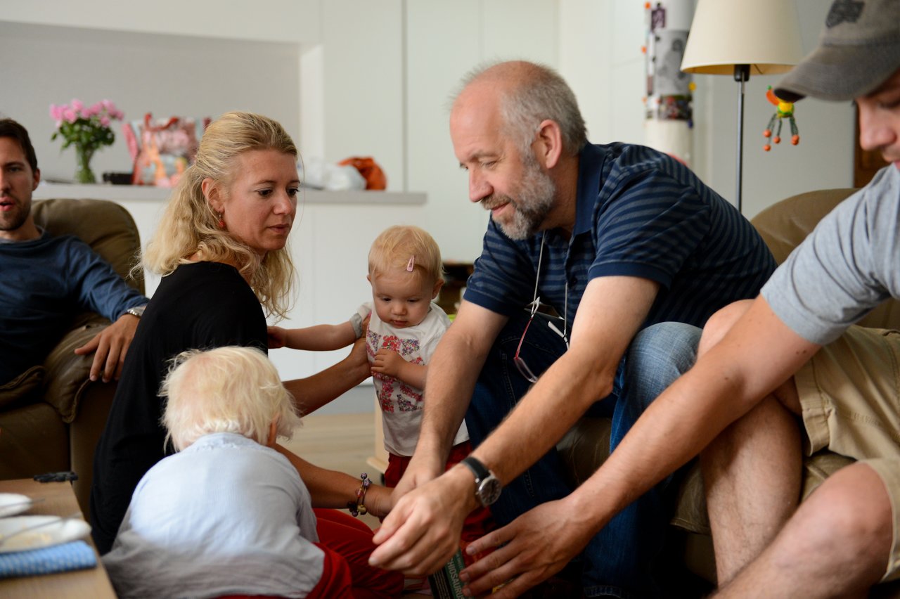 A family gathers in a living room, with adults interacting with two young children during a shared activity.