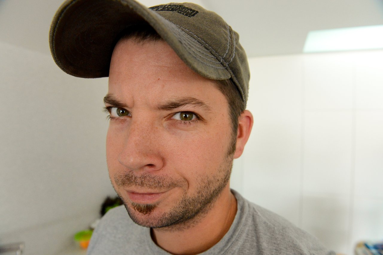 A man wearing a cap and gray shirt looks directly into the camera with a slightly raised eyebrow.