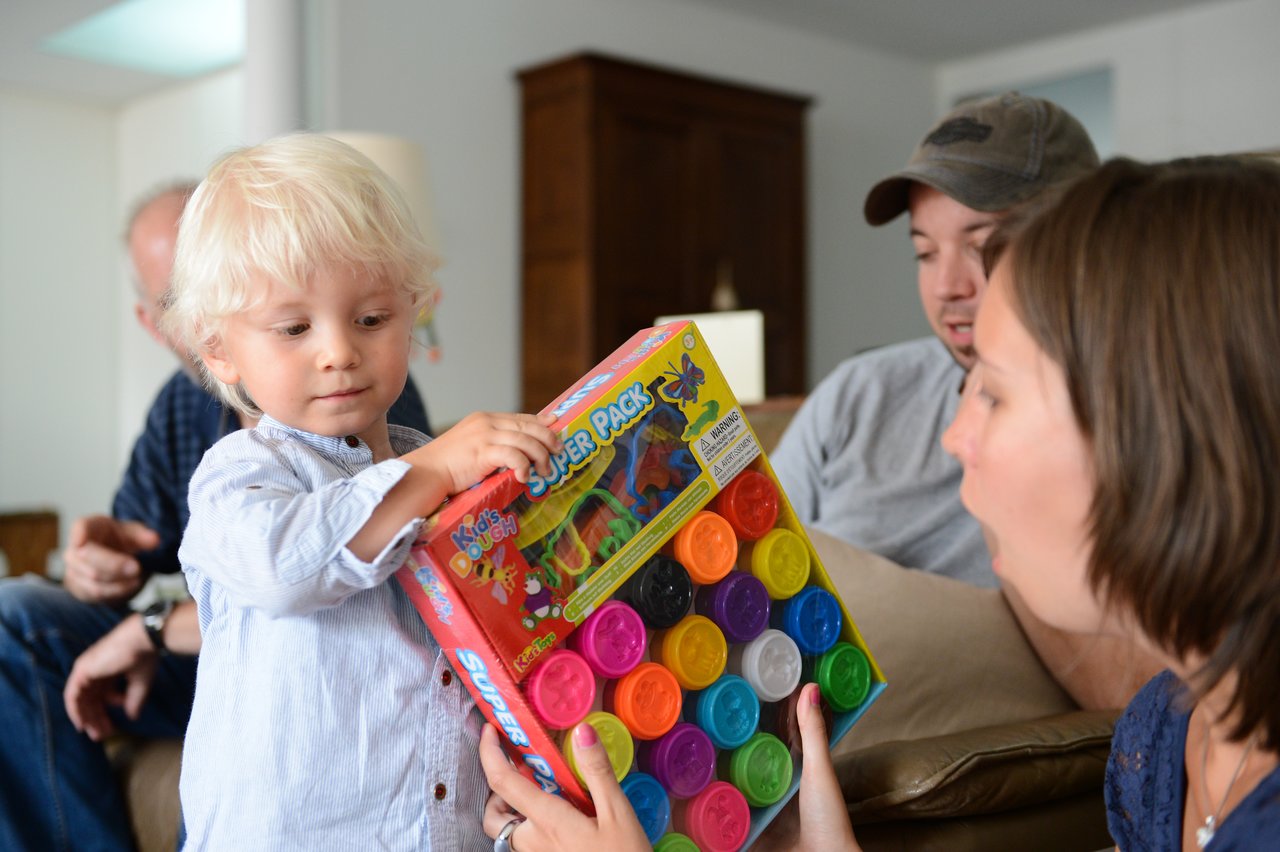 A young child holds a colorful playdough set while an adult reacts with excitement.