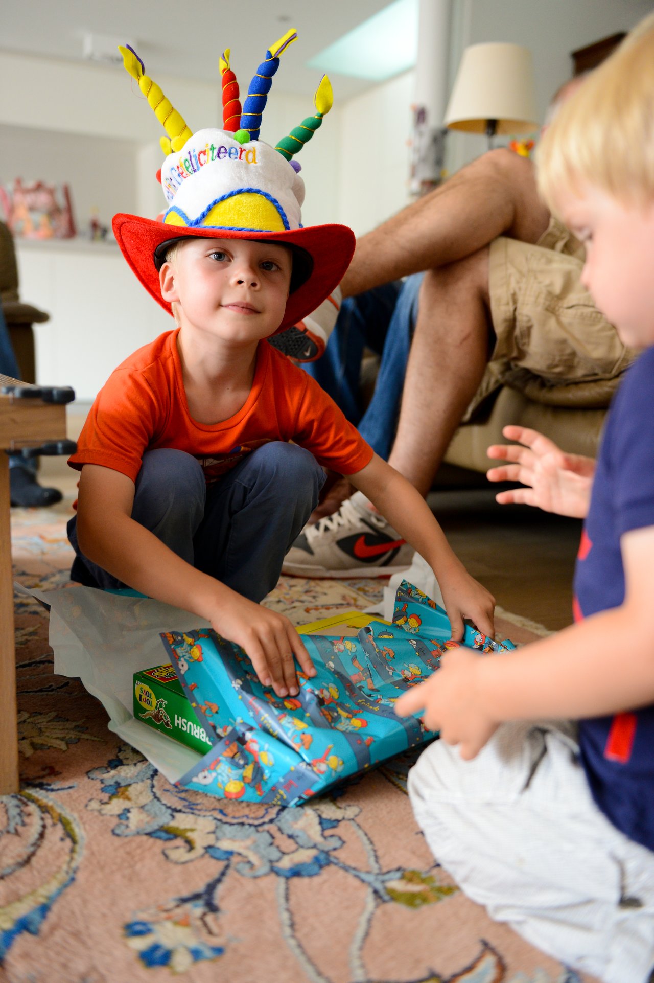 A young child wearing a festive birthday hat opens a wrapped present while another child watches nearby.