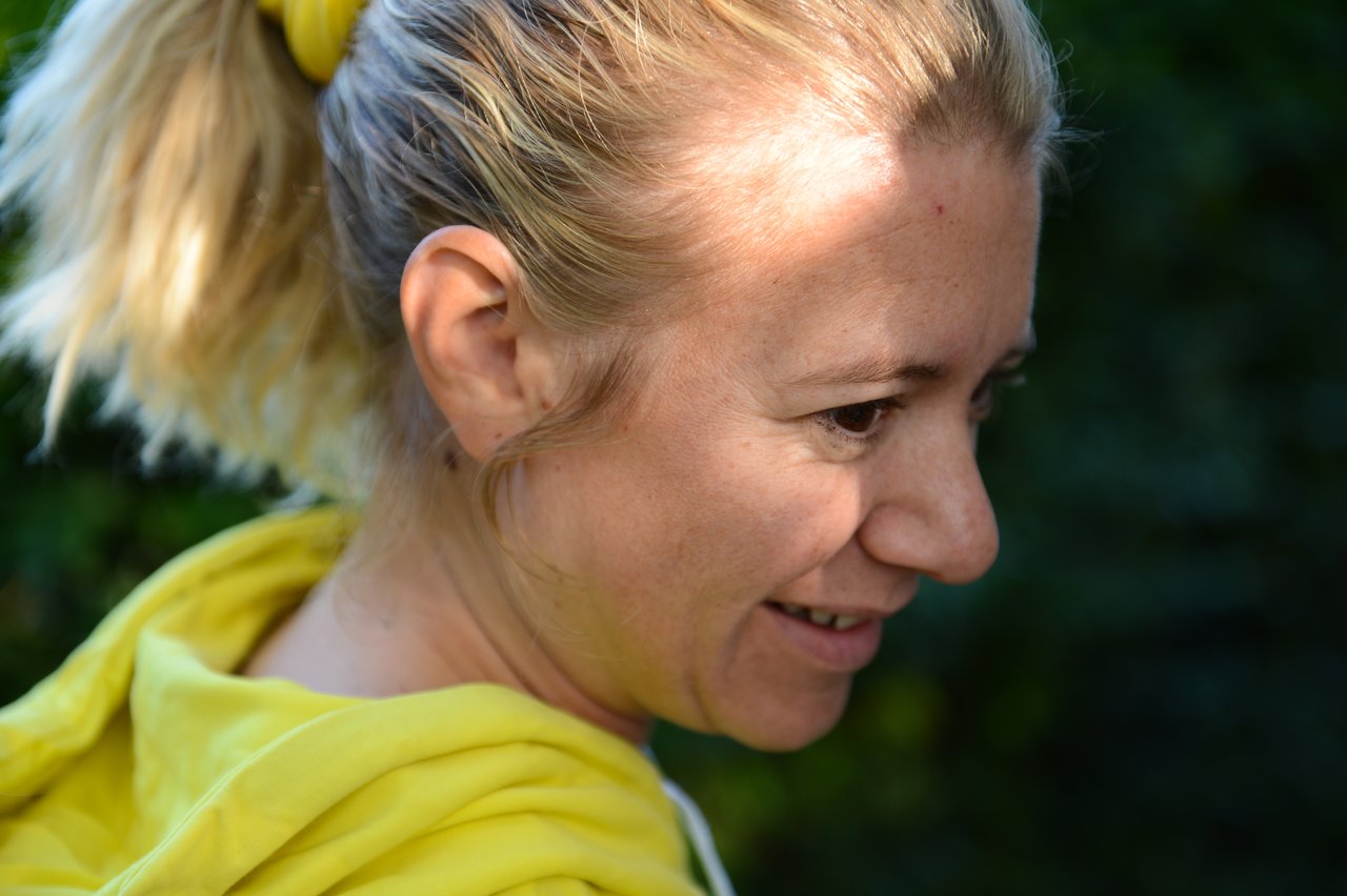 A woman in a yellow hoodie smiles while looking to the side, with sunlight on her face.