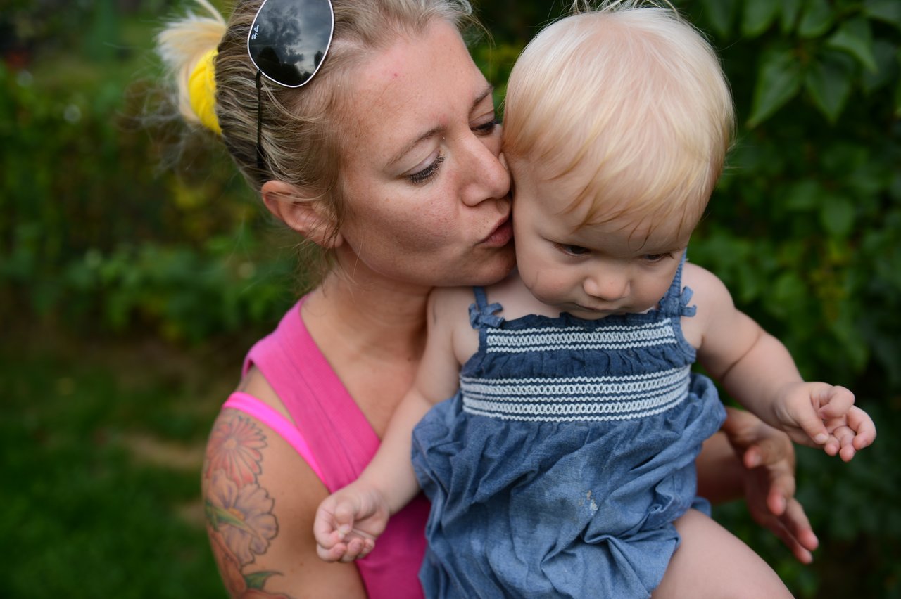 A woman in a pink top kisses a baby on the cheek while holding them outdoors.