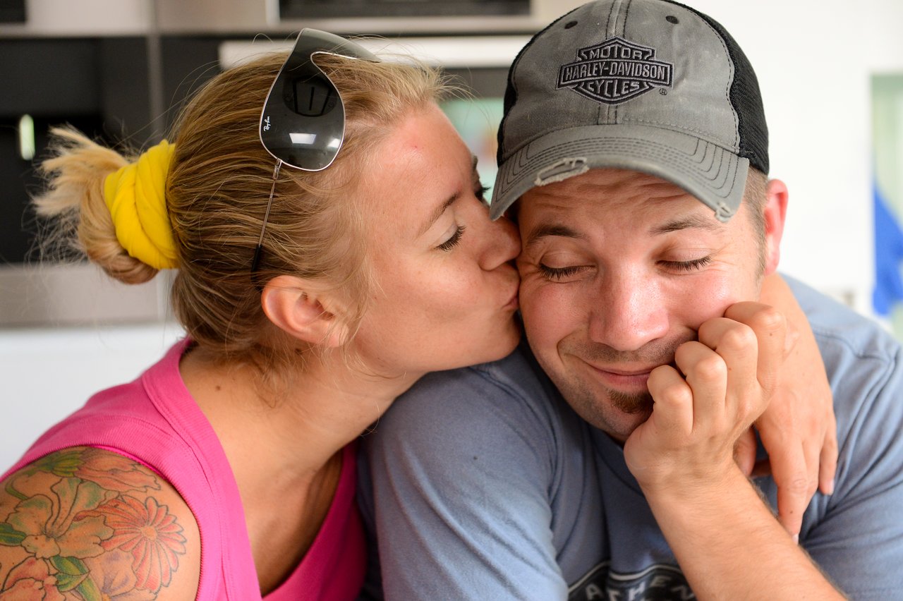 A woman in a pink top kisses a smiling man on the cheek as he rests his face on his hand.