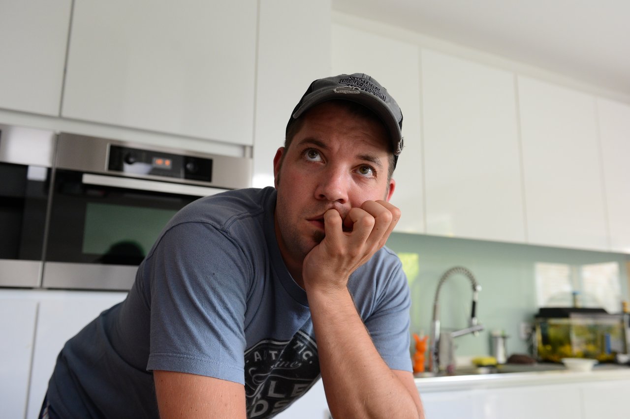 A man in a gray shirt and cap leans on a kitchen counter, resting his chin on his hand.