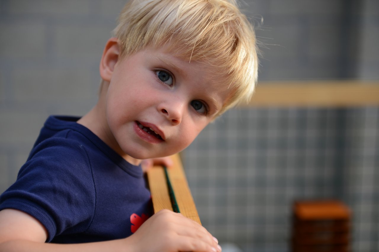 A young child with blonde hair leans on a wooden fence, looking curiously at the camera.