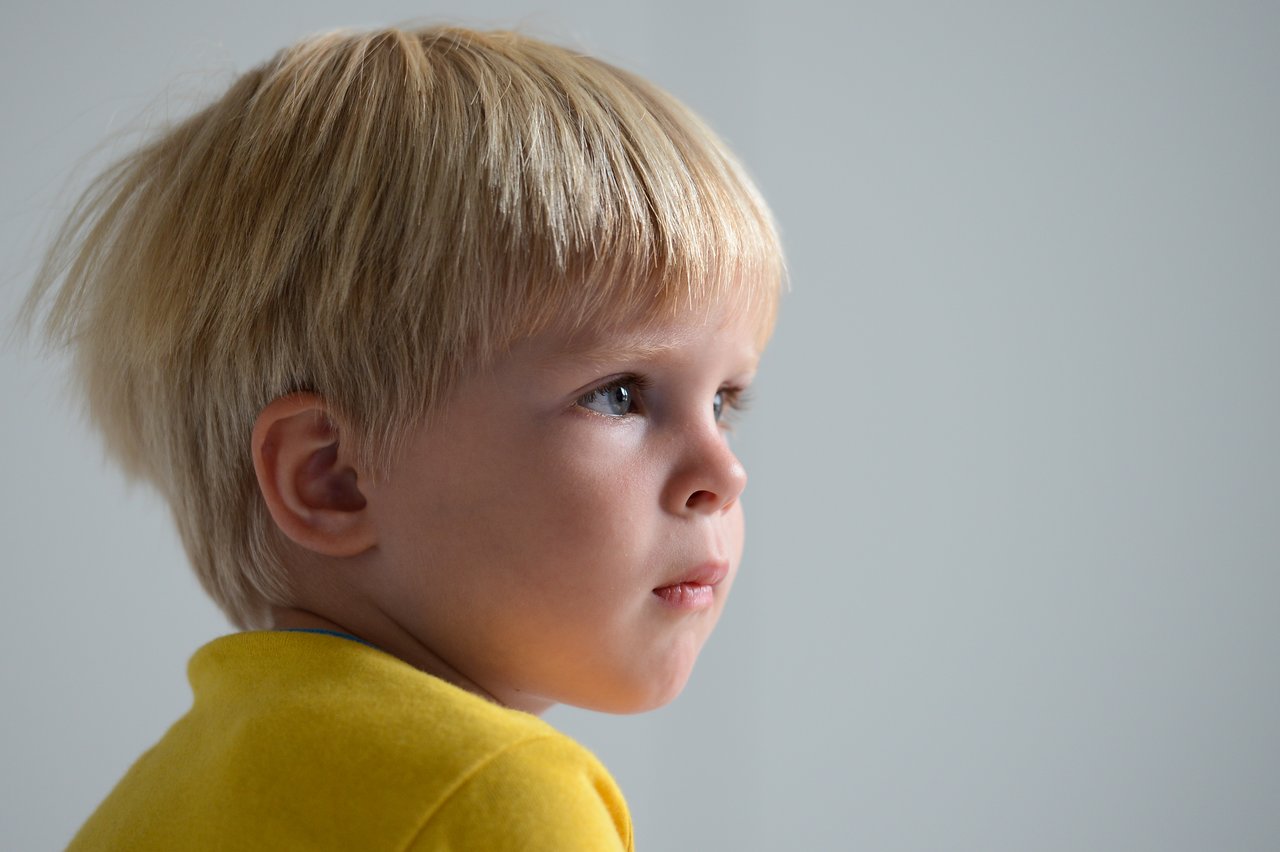 A young child with blond hair and a yellow shirt watches something intently, appearing focused.