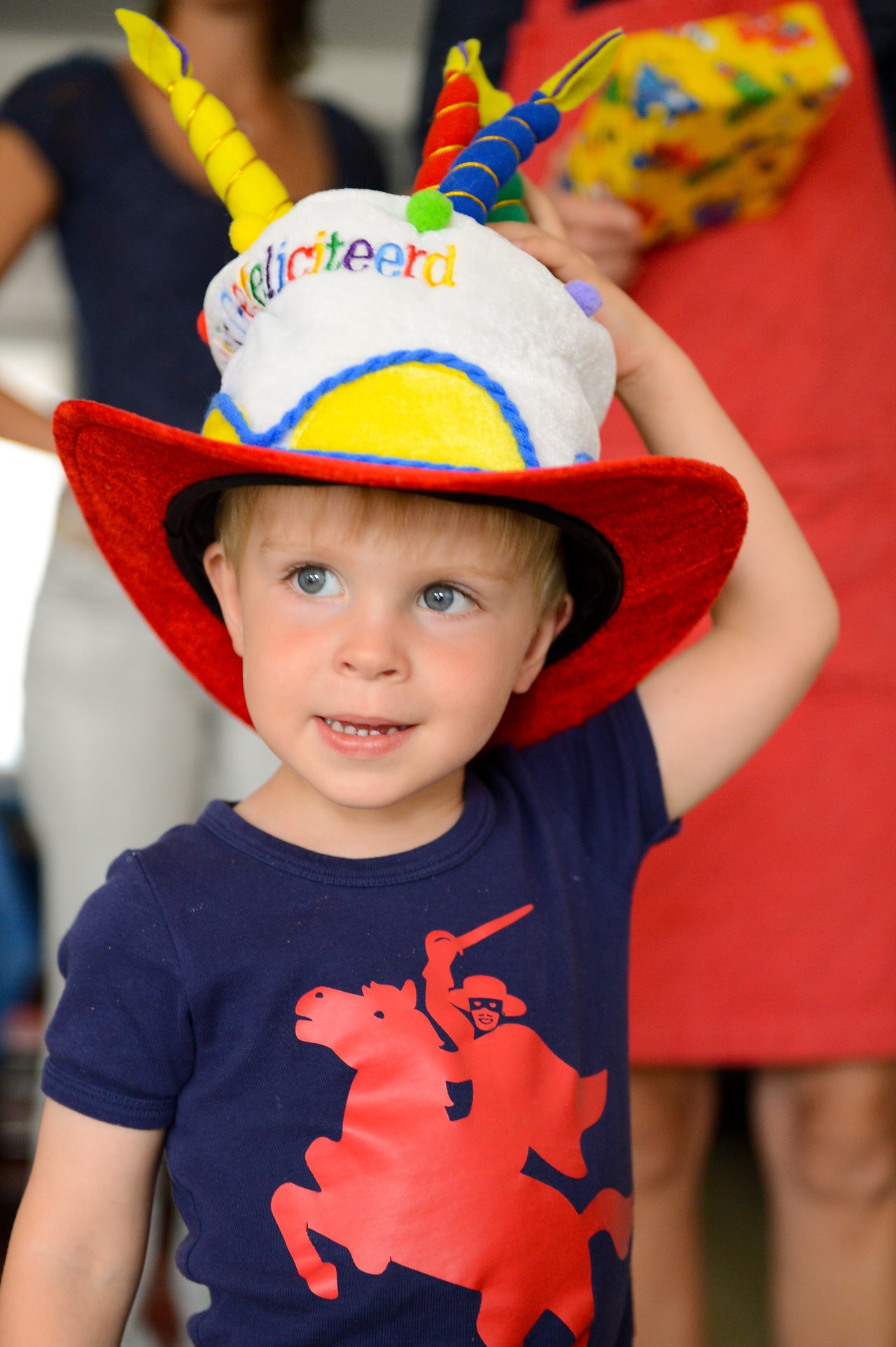 A young child wearing a colorful birthday hat and red cowboy-style brim, adjusting it with one hand.