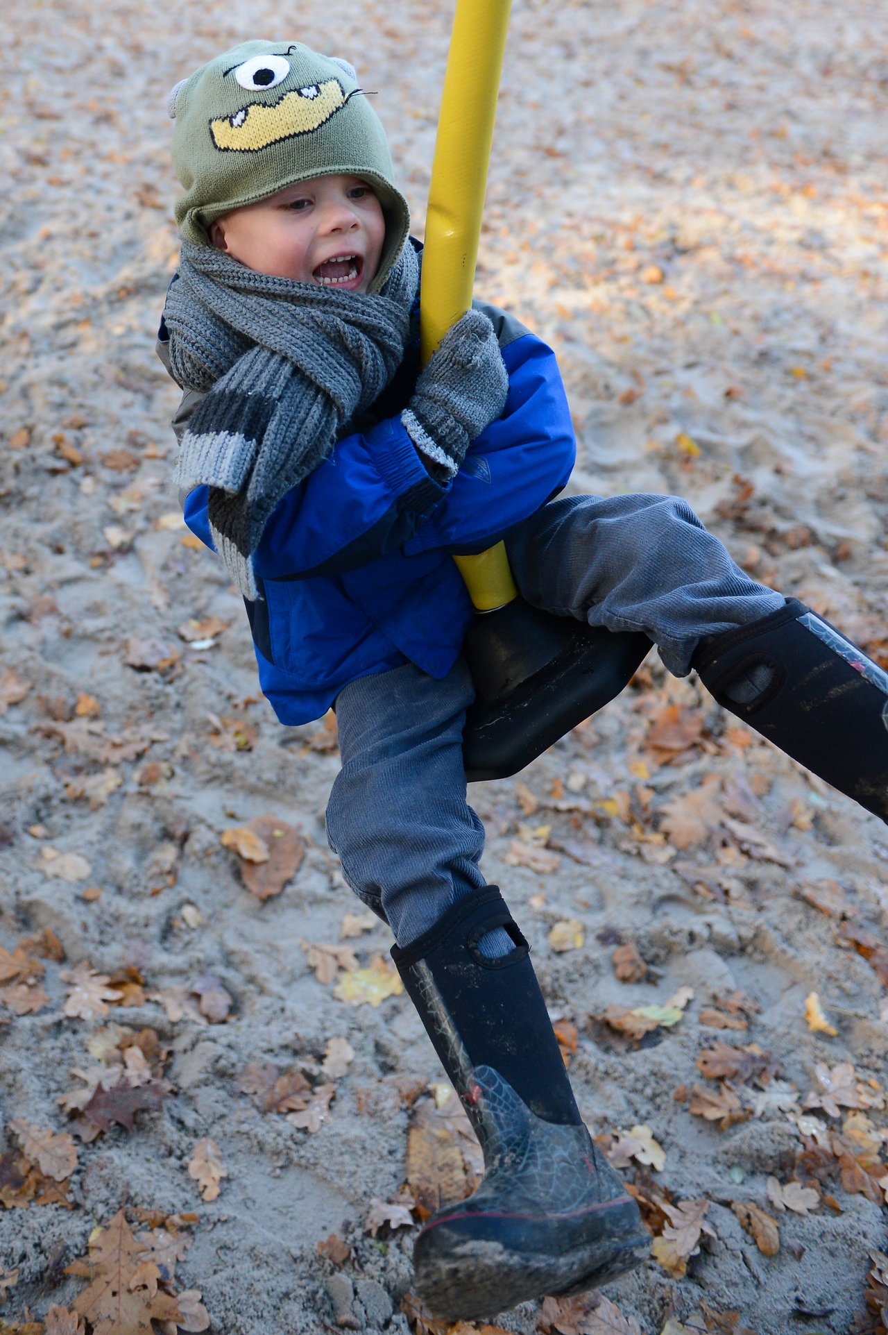 A child in a blue jacket and hat holds onto a playground swing, smiling while playing in the sand.