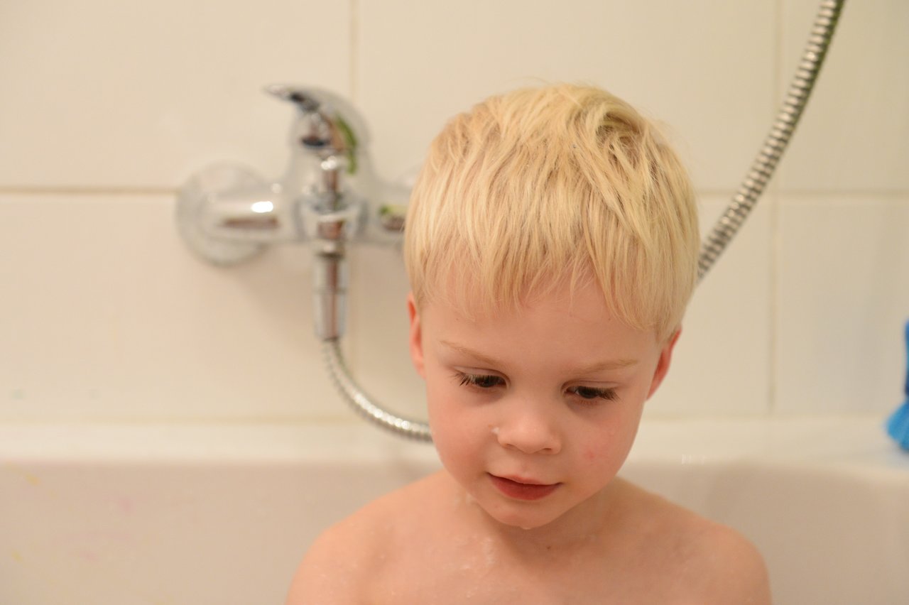 A young child with wet hair sits in a bathtub, looking down with a calm expression.
