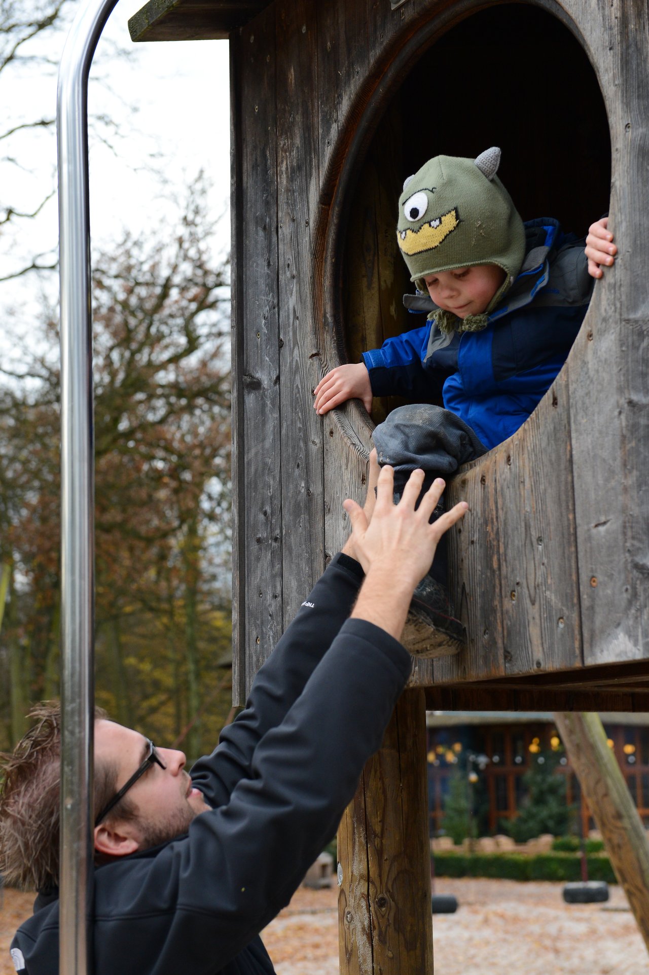 A father reaches up to help his child climb down from a wooden play structure at a playground.