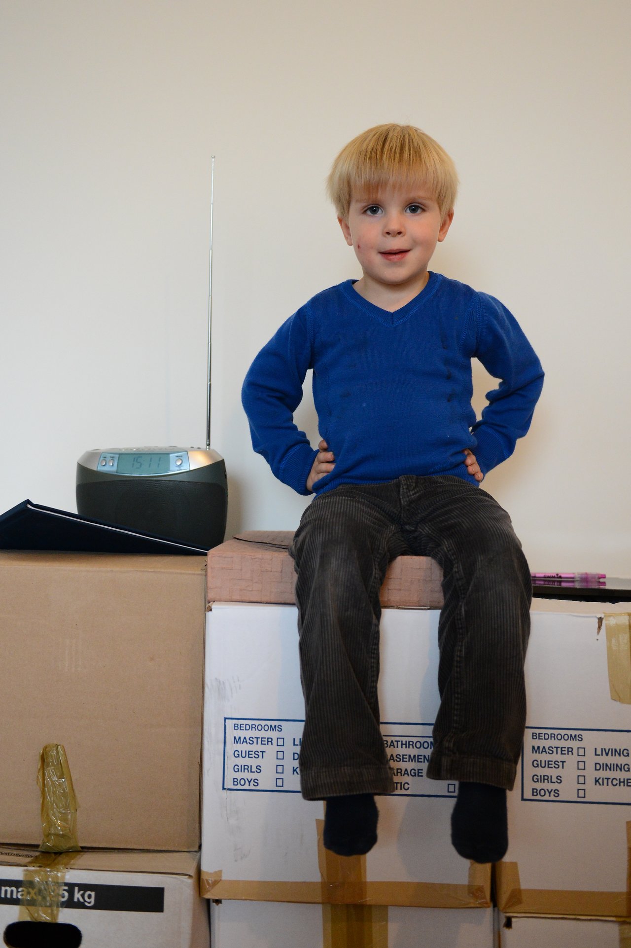 A young child in a blue sweater sits on stacked boxes with hands on hips, next to a radio.