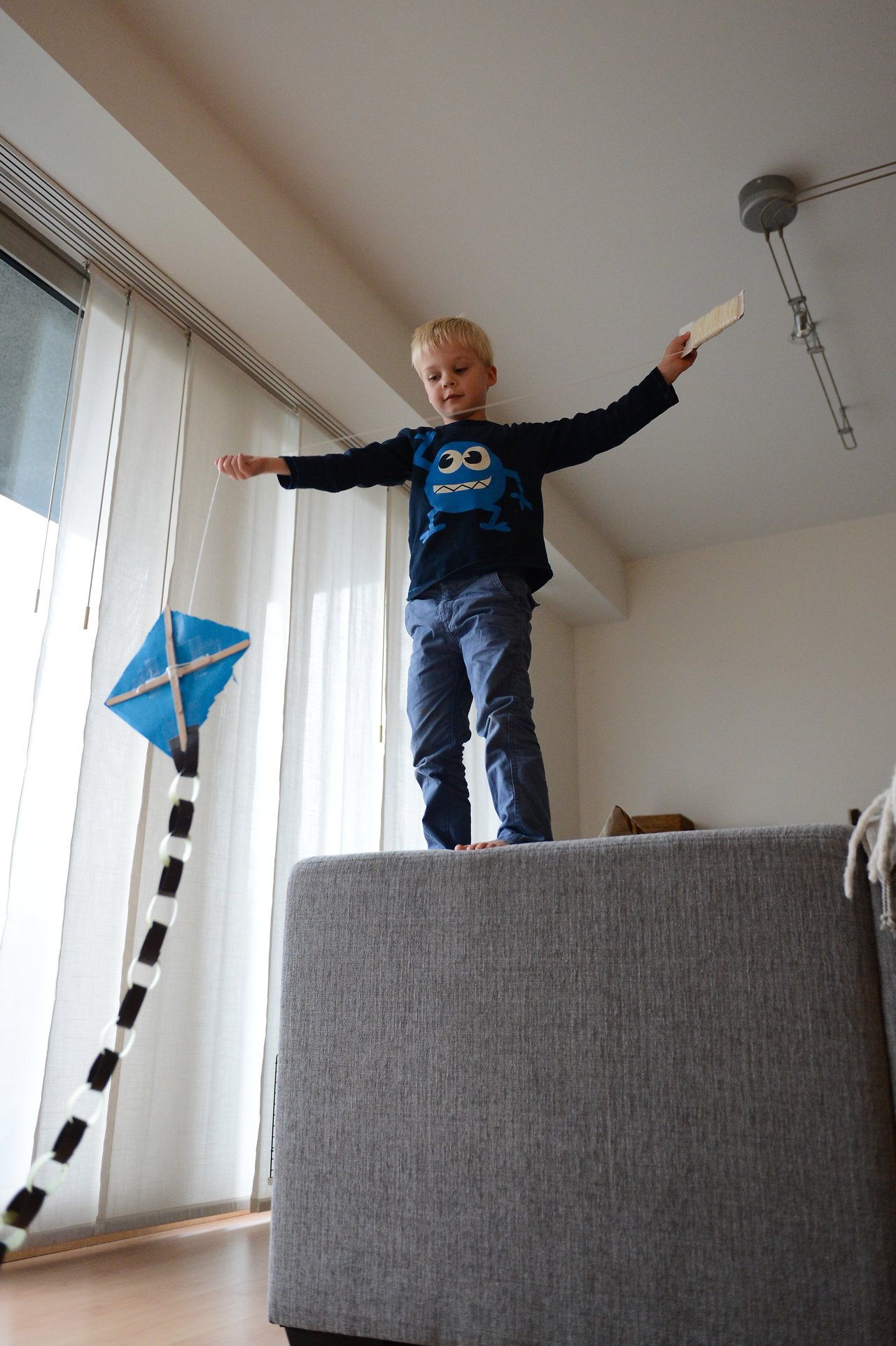 A young child stands on a couch, holding a string to fly a small blue kite indoors.
