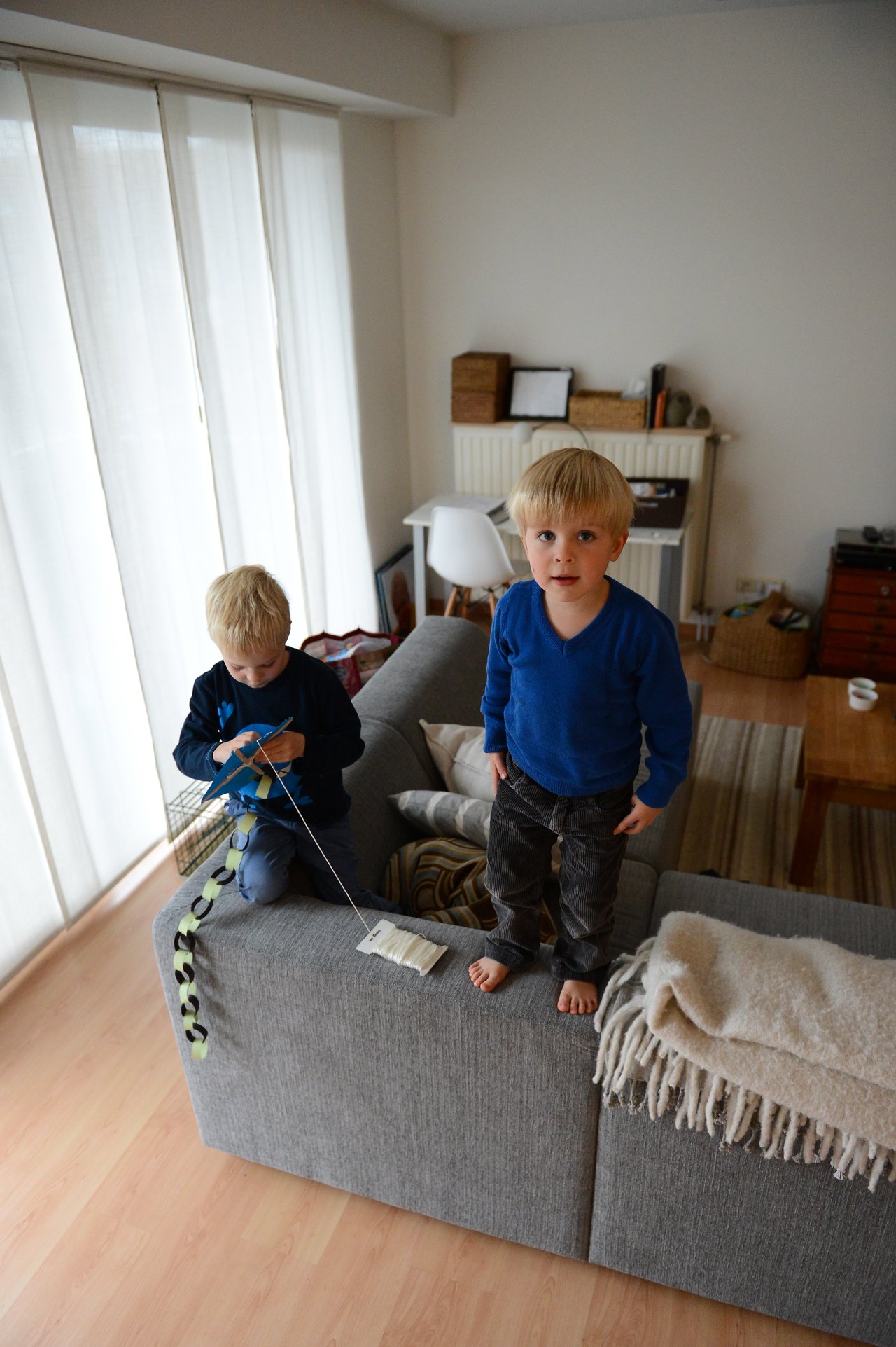 Two young children in blue shirts play with a kite indoors, one standing on the couch and the other holding the string.