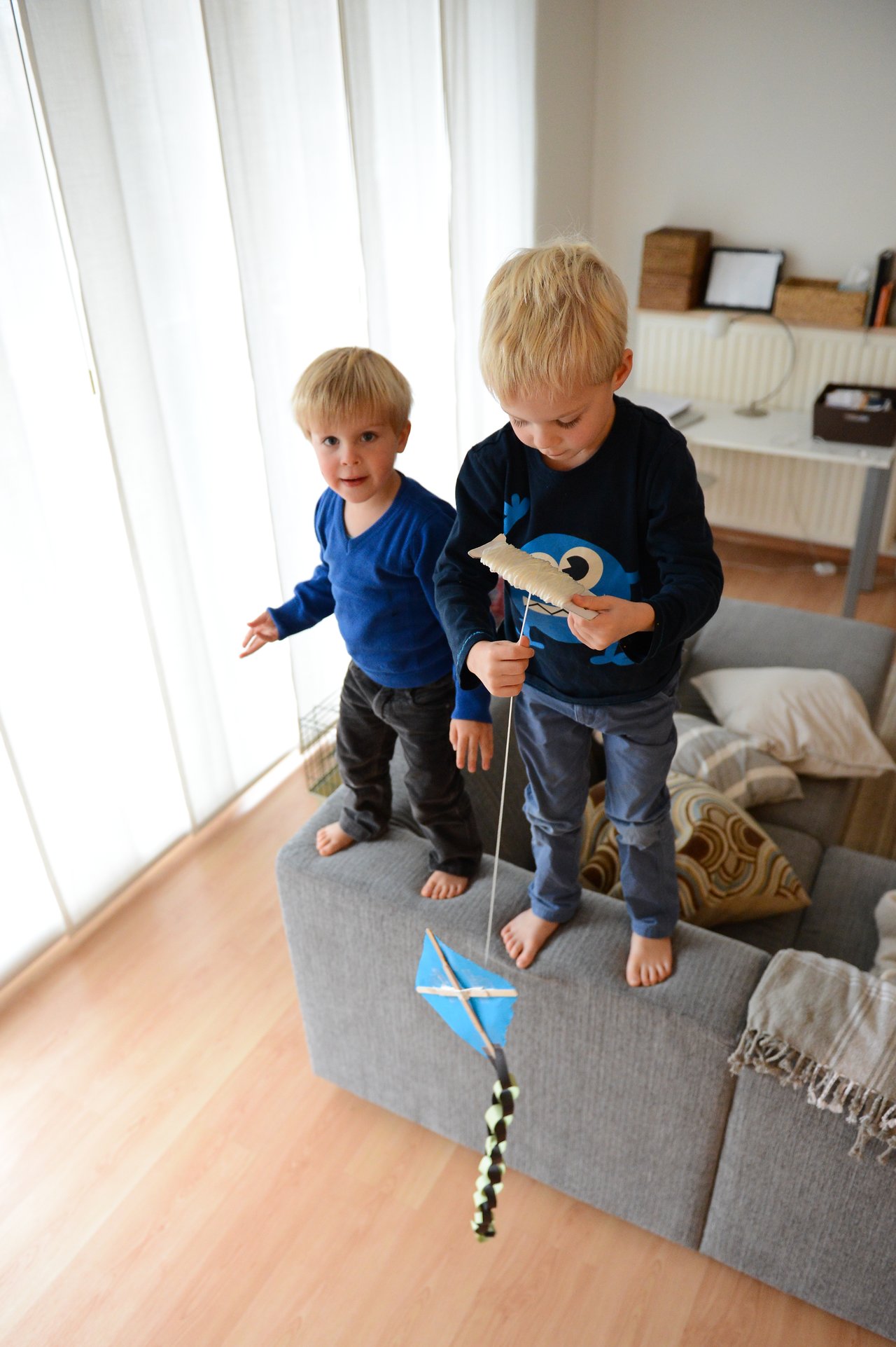 Two young children stand on a couch, holding a small blue kite with a string and tail.