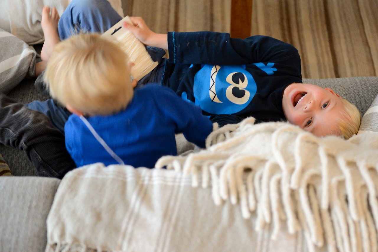 Two young children play and laugh on a couch, one holding a book while the other leans back, smiling.