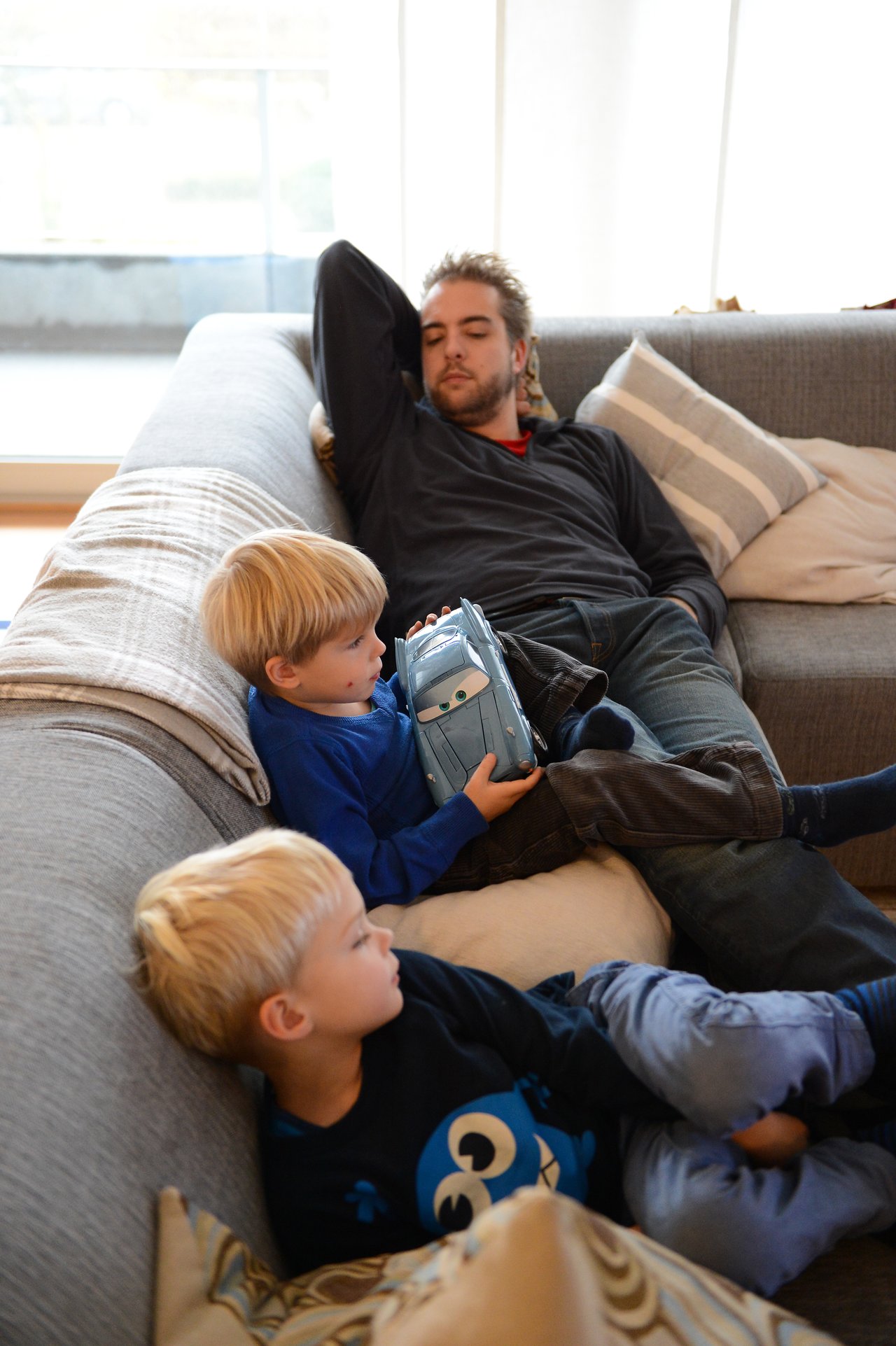 A man sleeps on a couch while two young children sit nearby, one holding a toy car.