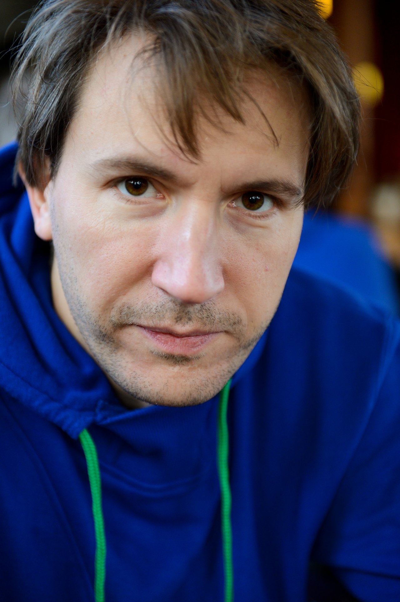 A man in a blue hoodie with green drawstrings looks directly at the camera with a neutral expression.