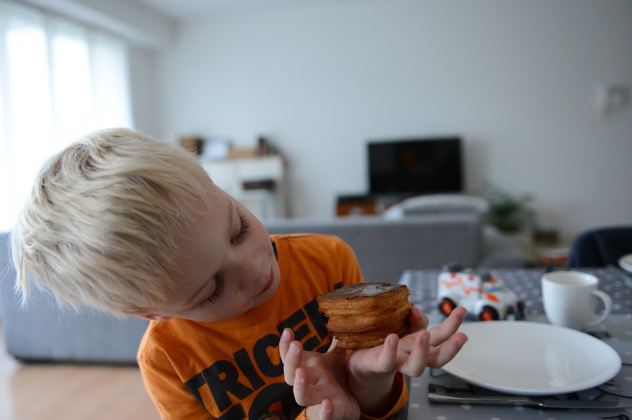 A child in an orange shirt holds a chocolate-topped pastry while sitting at a table set for lunch.