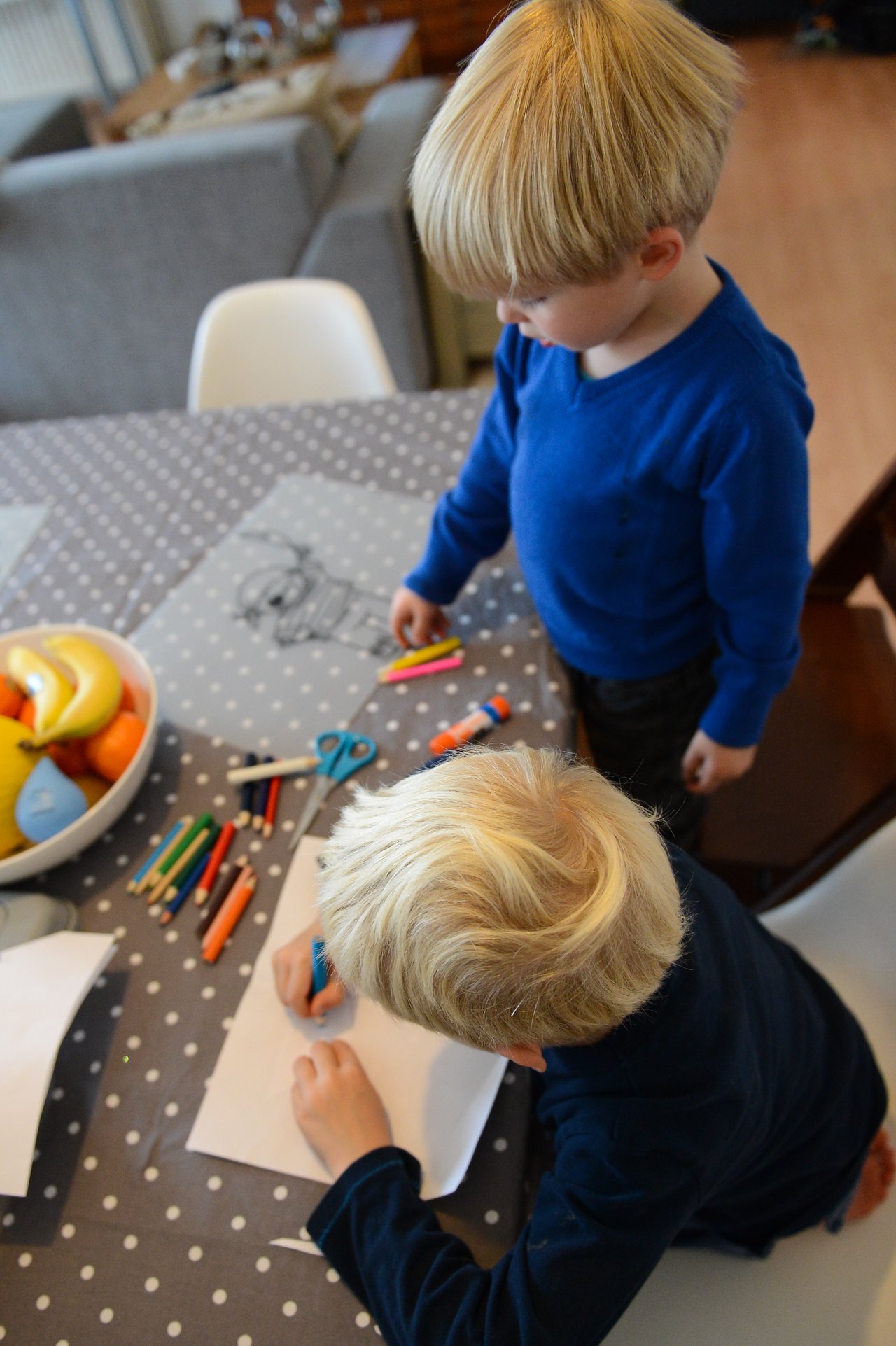 Two children at a table, one drawing on paper with a blue marker while the other watches.