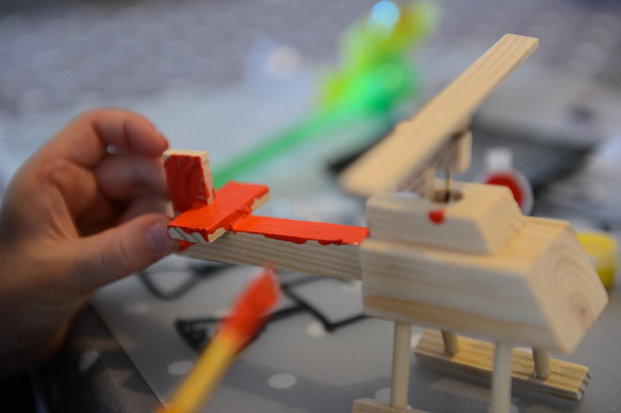 A person paints the tail of a small wooden helicopter model with red paint.