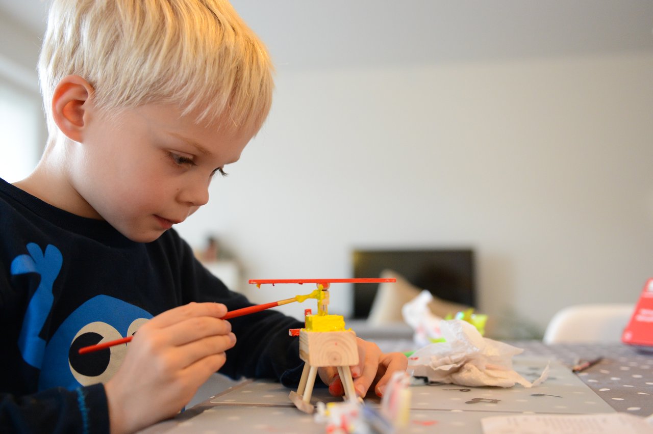 A child paints a small wooden helicopter with a red brush while sitting at a table.