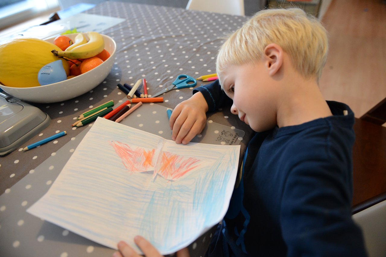 A child leans on a table, looking at a handmade butterfly pop-up card with colored pencils scattered nearby.