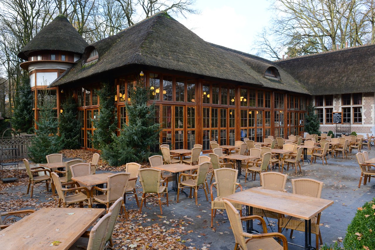 Outdoor seating area of a café with wooden tables and wicker chairs, surrounded by trees and a thatched-roof building.