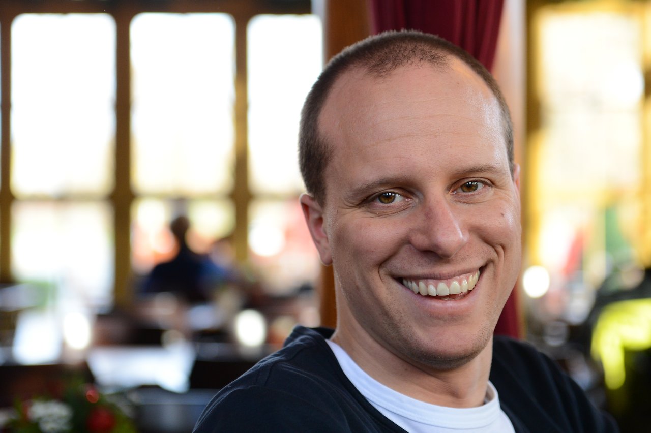 A man with a shaved head and a big smile looks directly at the camera in a well-lit indoor setting.