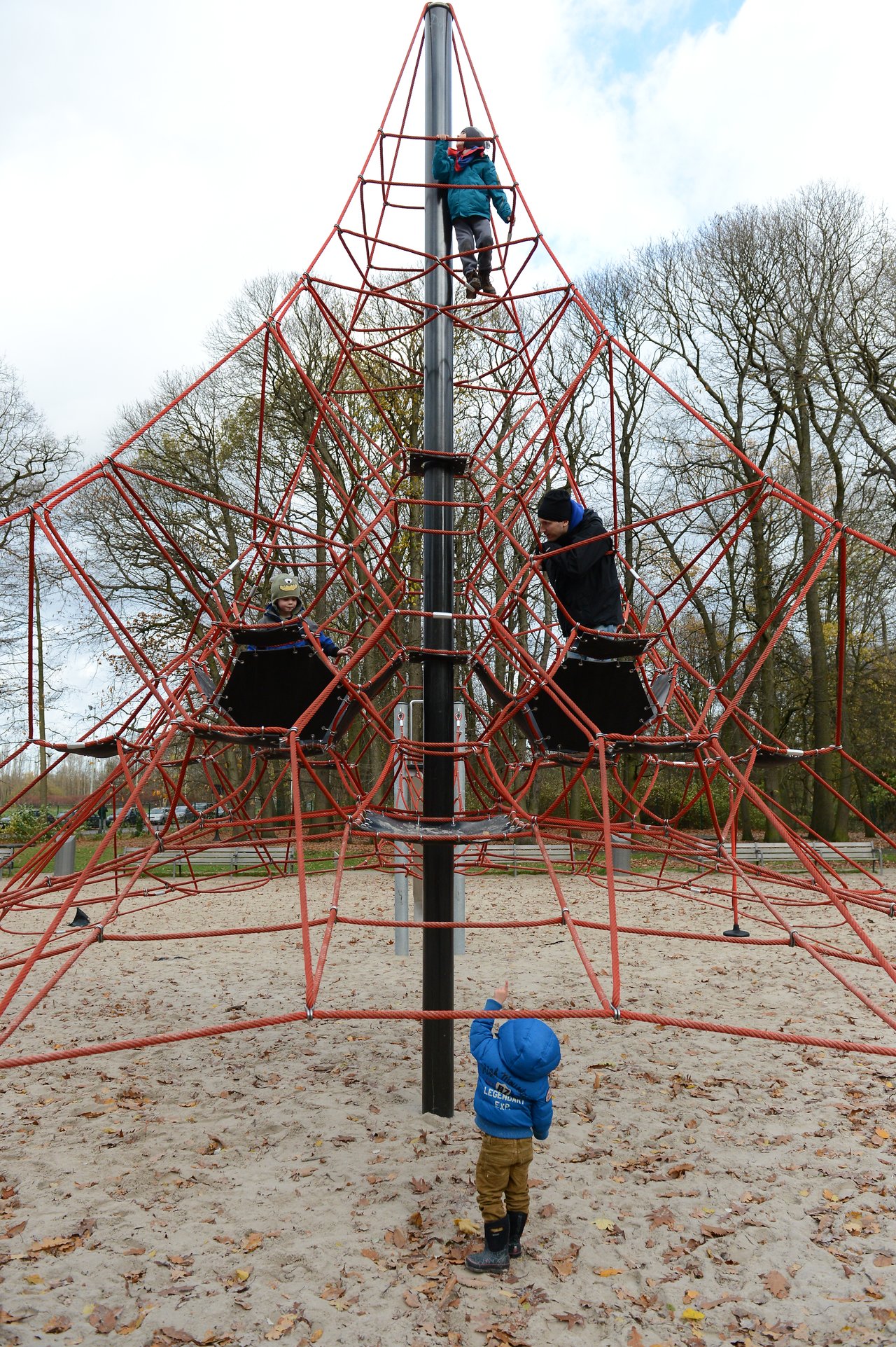 Children climb a tall rope playground structure while a younger child in a blue jacket watches from the ground.