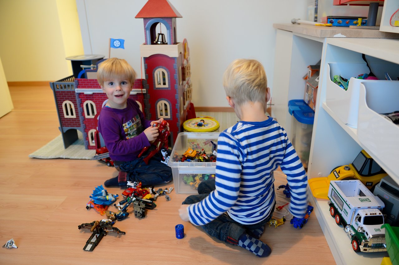 Two young children sit on the floor, playing with action figures and toy vehicles near a large toy castle.