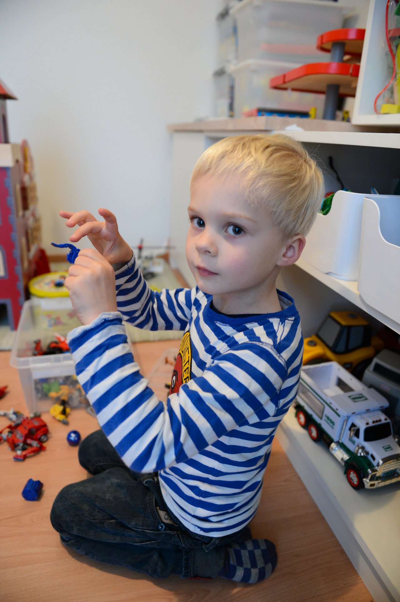 A young child in a striped shirt sits on the floor, holding a small toy while surrounded by other toys.