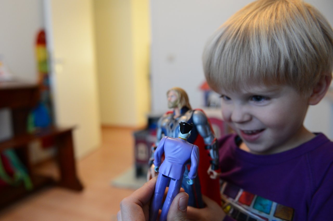 A smiling child in a purple shirt plays with action figures, holding one while another is in the background.