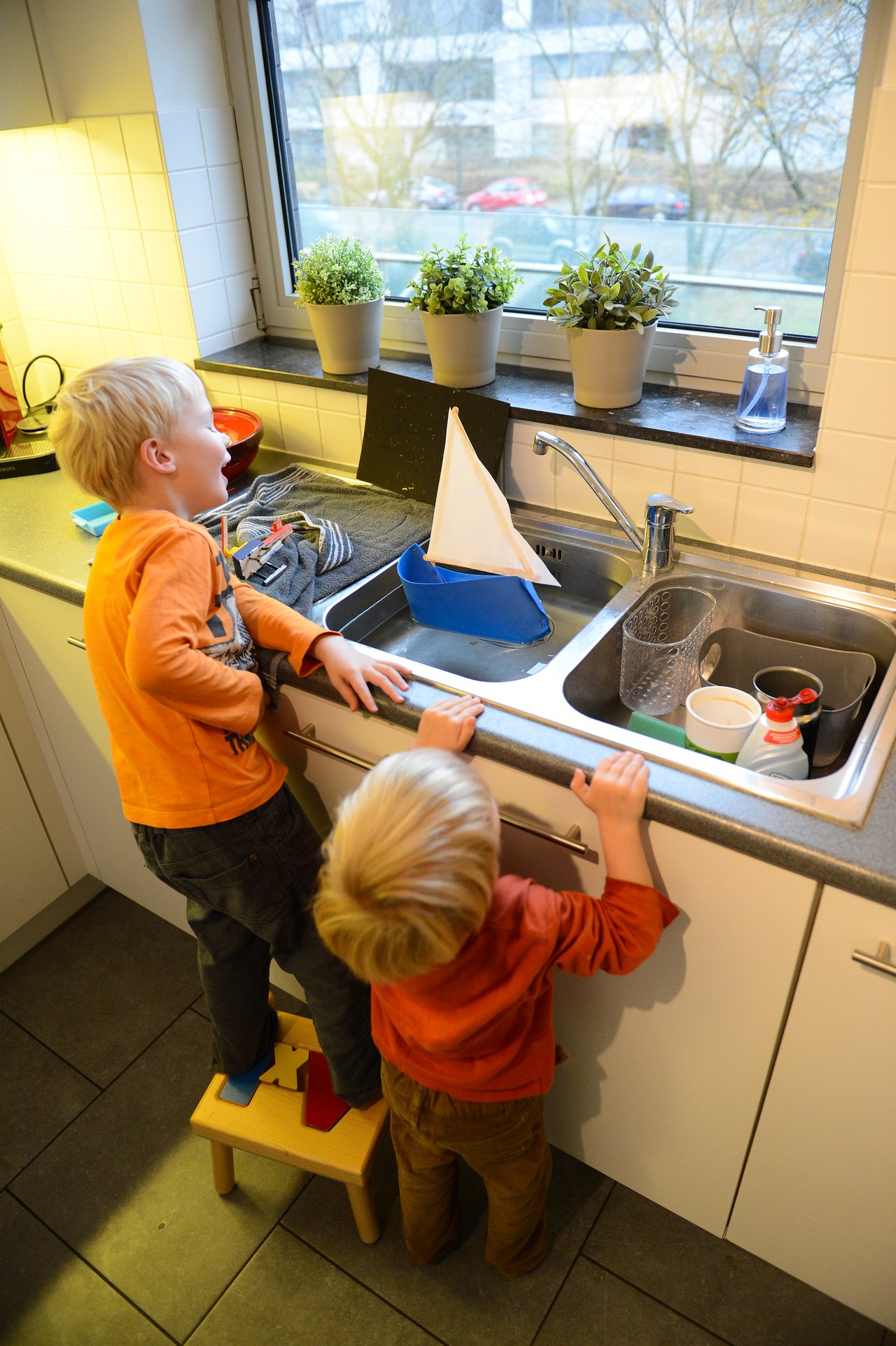 Two children in an orange outfit watch a small homemade sailboat floating in a kitchen sink.