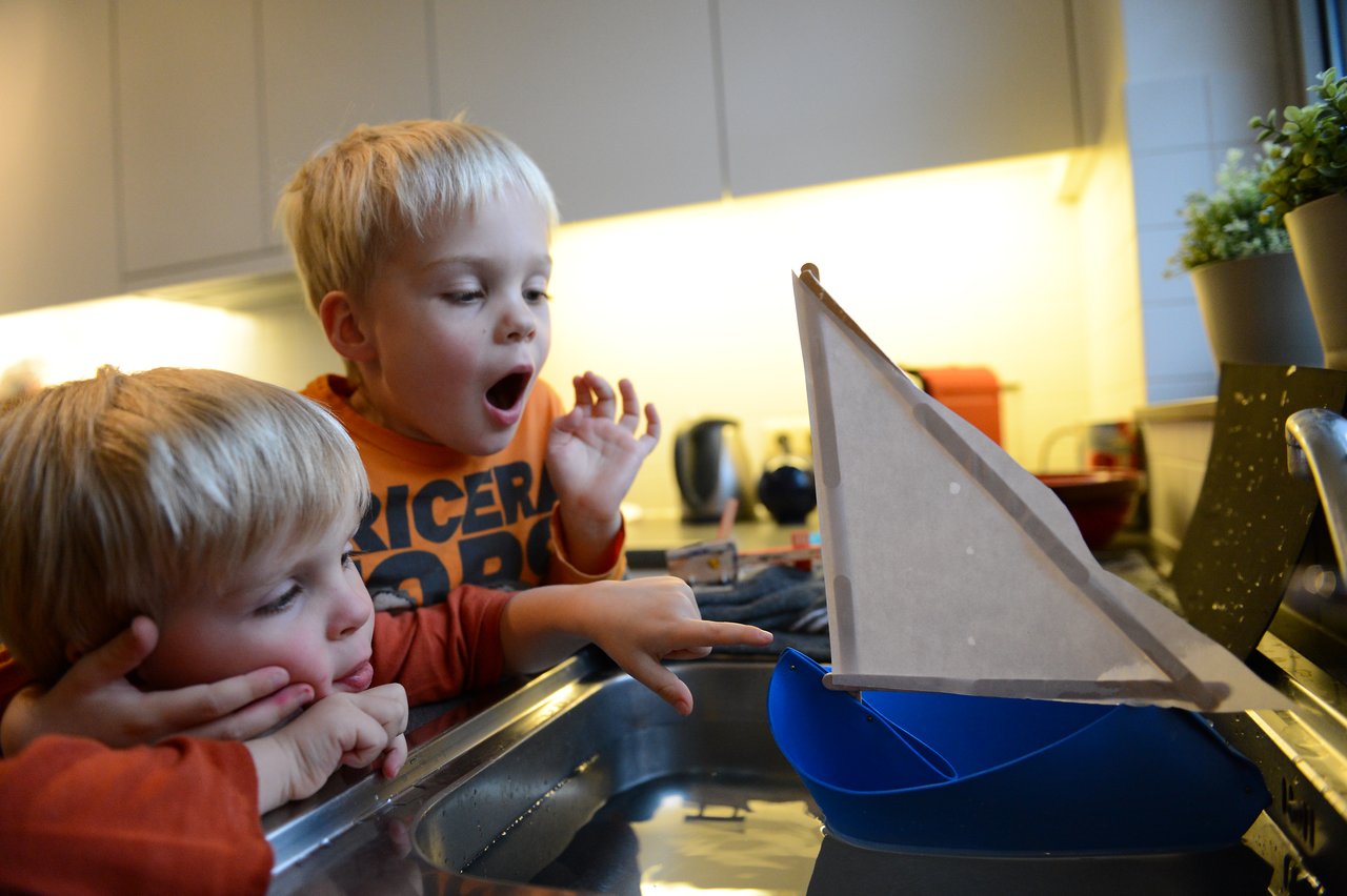Two young children watch excitedly as a homemade sailboat floats in a kitchen sink.