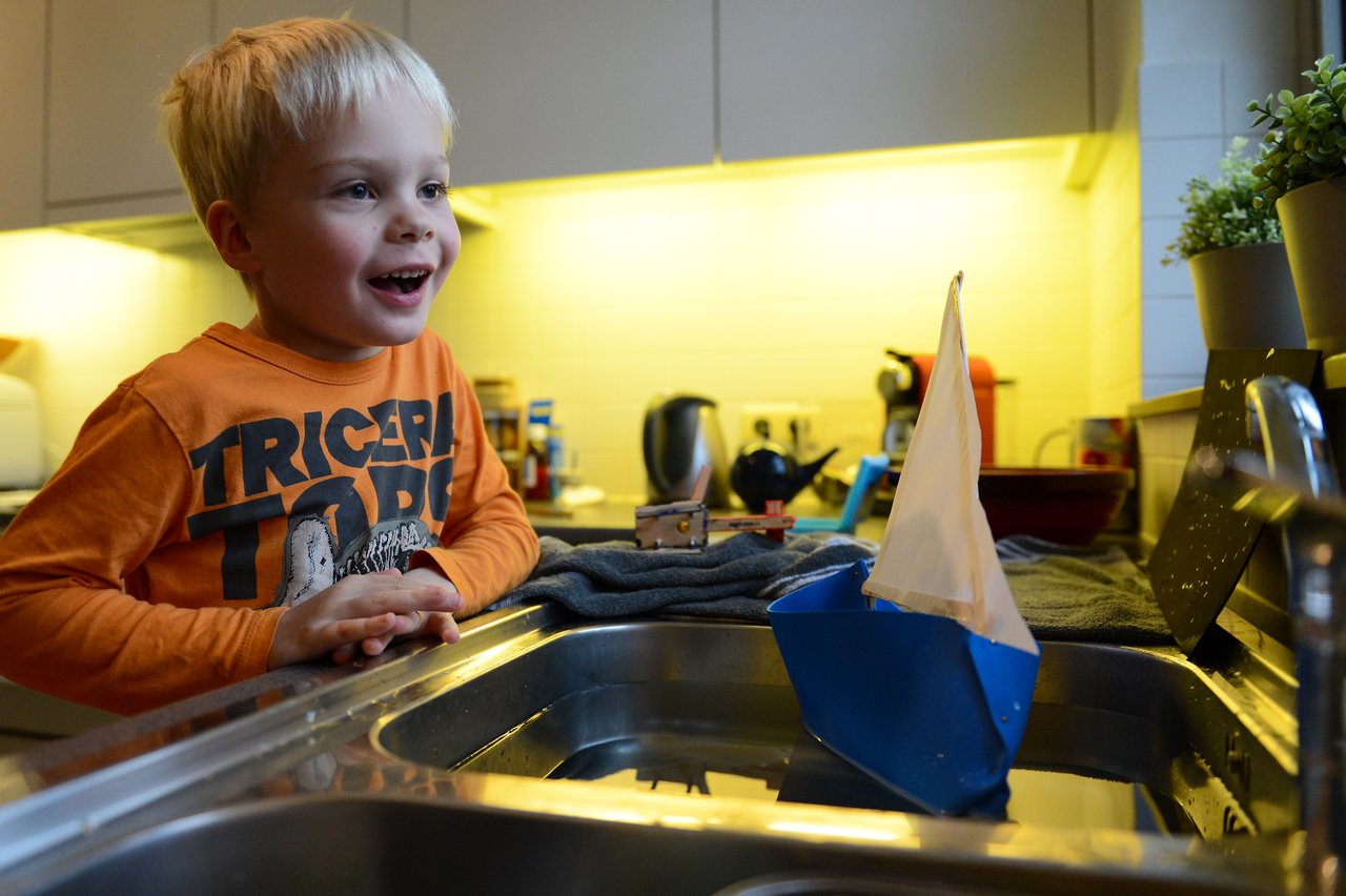 A young child excitedly watches a homemade sailboat float in a kitchen sink filled with water.