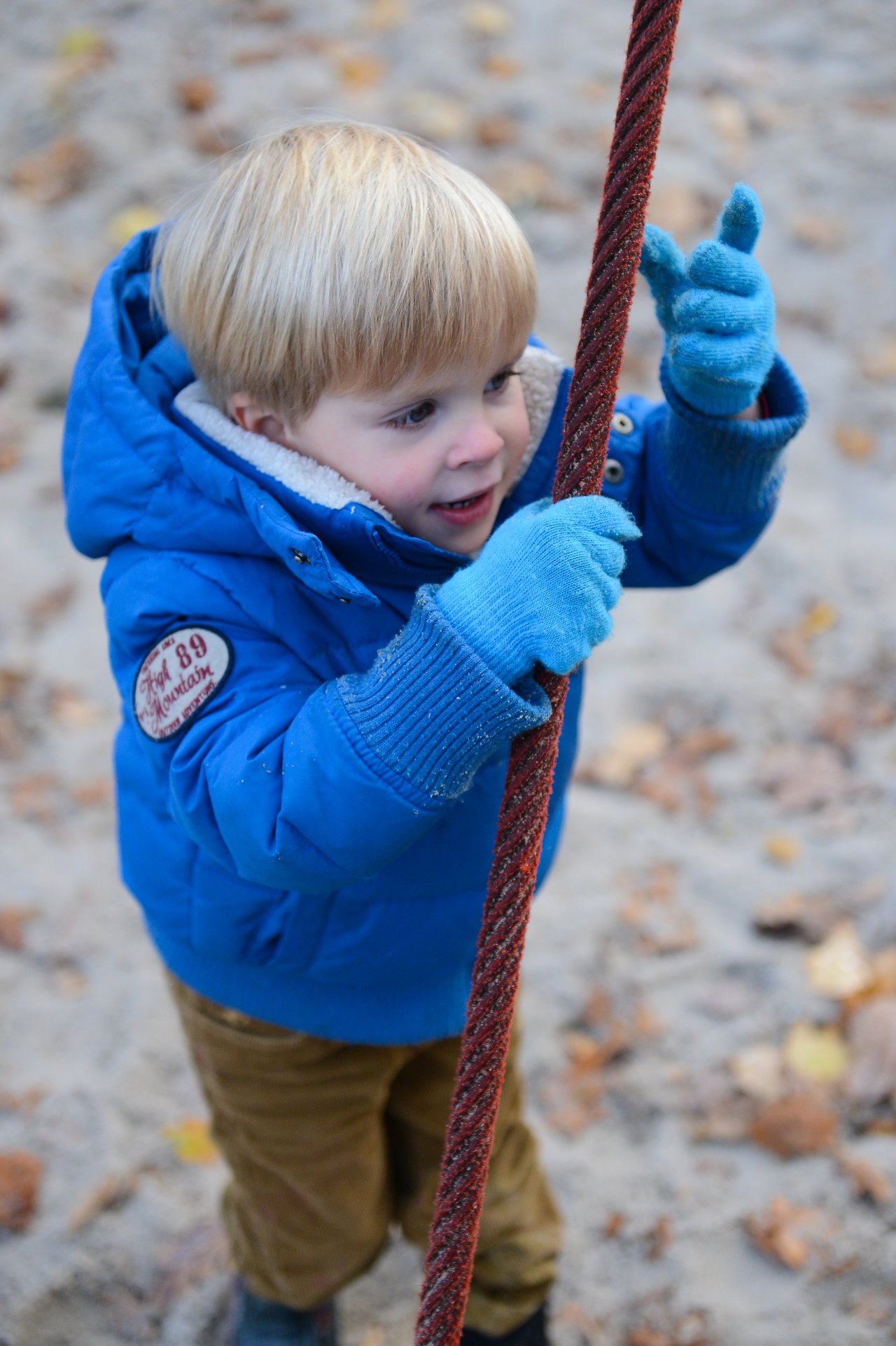 A young child in a blue jacket and gloves holds onto a rope at a playground.