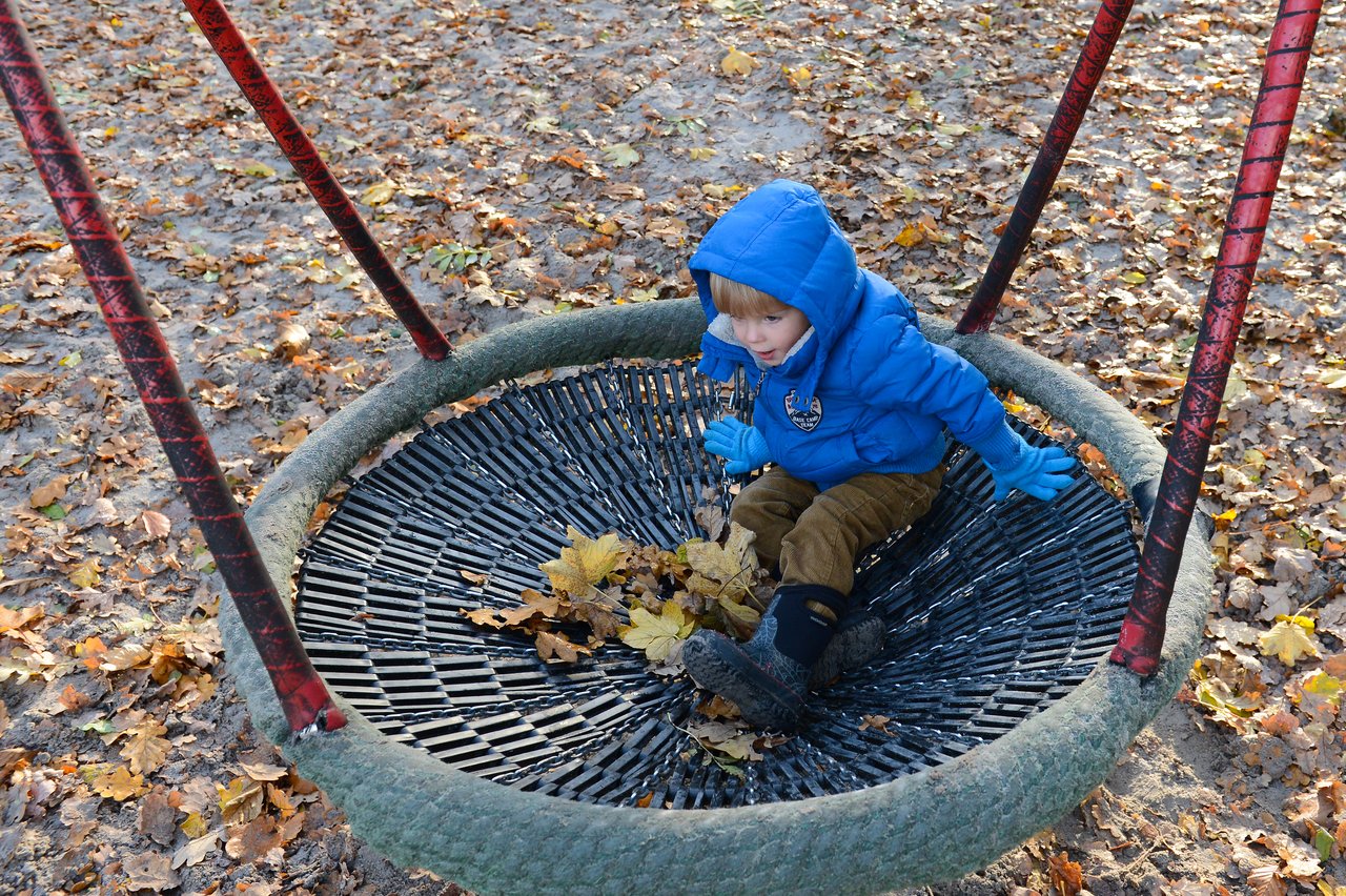 A child in a blue jacket sits on a large swing at a playground, surrounded by fallen autumn leaves.