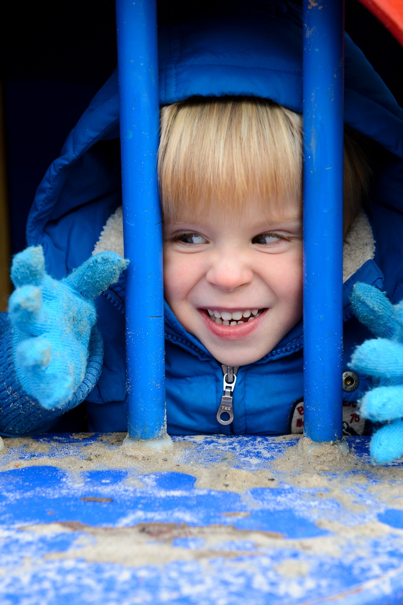 A child in a blue jacket and gloves smiles while peeking through blue playground bars.