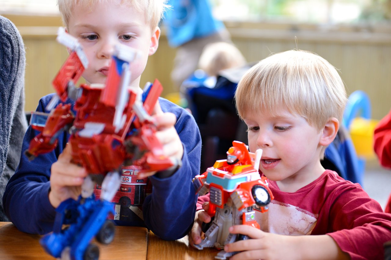 Two young boys sit at a table, playing with colorful Transformer action figures, focusing intently on their toys.